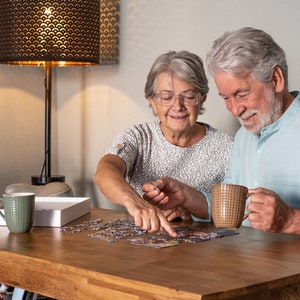 Może przedstawiać: Para siedzi przy stole i układa puzzle. Kobieta trzyma kawałek puzzli, a mężczyzna trzyma kubek. St&oacute;ł jest drewniany i ma jasnobrązowy kolor. W tle znajduje się lampa.