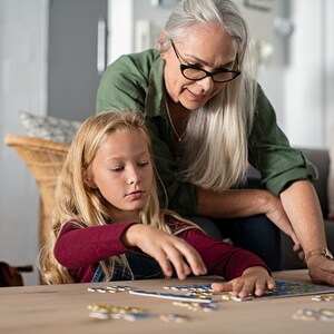 Może przedstawiać: Babcia i wnuczka wsp&oacute;lnie układają puzzle na drewnianym stole. Babcia ma na sobie zieloną koszulę i okulary, a wnuczka czerwoną koszulę.