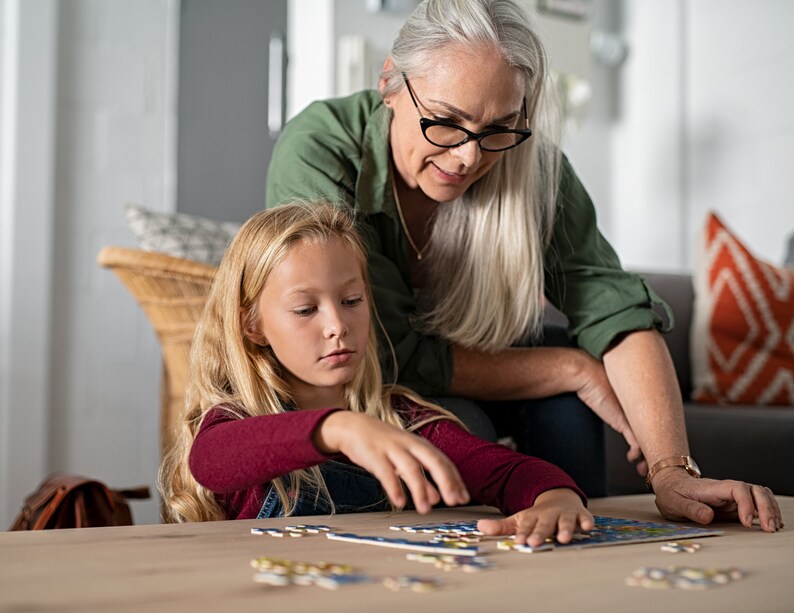 Może przedstawiać: Babcia i wnuczka wsp&oacute;lnie układają puzzle na drewnianym stole. Babcia ma na sobie zieloną koszulę i okulary, a wnuczka czerwoną koszulę.