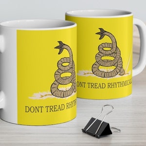 Op de afbeelding: Twee witte keramische mokken met een gele achtergrond en een bruin slangenontwerp. De mokken hebben de tekst "Don't Tread Rhythmically" erop.