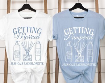 May include: Two white t-shirts with blue text and graphics. The left shirt says "Getting Married" and the right shirt says "Getting Pampered". Both shirts have a graphic of three bottles of spa products and say "Jessica's Bachelorette" at the bottom.