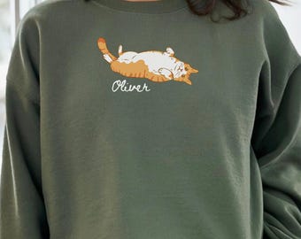 May include: Military green sweatshirt with a graphic of a cat lying on its back. The cat is orange and white, and the name "Oliver" is written below it in cursive. The sweatshirt is a solid color and has long sleeves.