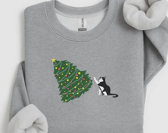 Può includere: Una felpa girocollo grigio chiaro con una stampa grafica di un albero di Natale verde decorato con ornamenti gialli e rossi e una stella gialla in cima, con un gatto bianco e nero che allunga una zampa anteriore verso l'albero.