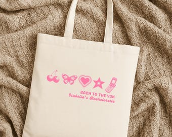 Puede incluir: Una bolsa de tela color crema con un estampado gráfico rosa de cerezas, una mariposa, un corazón, una estrella y un teléfono plegable. Debajo de los iconos, el texto dice "BACH TO THE Y2K" y "Isabella's Bachelorette" en fuente rosa.