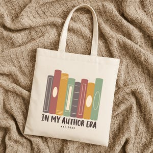 May include: Cream-colored tote bag with a row of colorful book illustrations. The text "IN MY AUTHOR ERA est. 2025" is printed below the books. The bag has two handles and is made of a sturdy fabric.