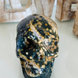 Puede incluir: Un cráneo negro, blanco y marrón hecho de jaspe oceánico pulido. El cráneo tiene una talla detallada de los rasgos faciales y los dientes.
