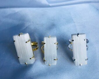 May include: Three selenite crystal rings with gold and silver bands. The rings are on a light blue fabric background.