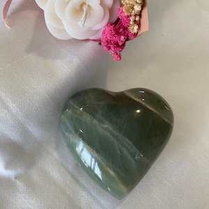 Könnte beinhalten: Ein grüner herzförmiger Stein mit weißen und braunen Streifen. Der Stein ist poliert und glatt.