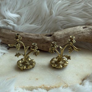 May include: A pair of brass candle holders, each featuring a circular base with a floral petal design. Two curved stems rise from the centre, branching out into two individual candle cups adorned with sculpted leaves and small flower accents.