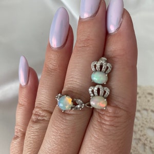 May include: A silver ring with two opal stones set in crown-shaped settings. The ring is on a finger with light purple nail polish.