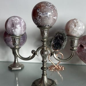 May include: A silver candelabra with three arms, each holding a spherical, patterned stone. The stones are shades of pink, purple, and white. The candelabra is on a reflective surface.