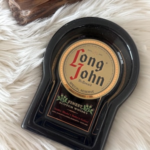 Op de afbeelding: Zwarte asbak in sleutelgatvorm, met een goud en rood label met de tekst "Long John McDonald Special Reserve Finest Scotch Whisky". De asbak staat op een witte, zachte ondergrond.