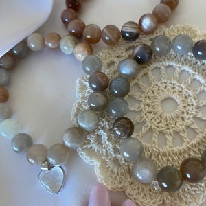 Könnte beinhalten: Drei Armbänder aus Sunstone-Perlen. Die Armbänder sind übereinander gestapelt und liegen auf einer weißen Häkelunterlage. Die Armbänder sind in verschiedenen Brauntönen, Grau und Weiß.