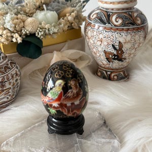 Pode incluir: Um objeto decorativo em forma de ovo preto e vermelho com um desenho pintado de dois pássaros em um galho. O objeto está sentado em um pequeno suporte preto e uma superfície de cristal branco.