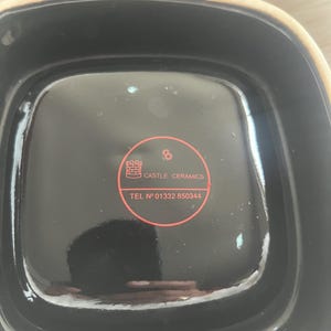 Puede incluir: Un cuenco cuadrado de cer&aacute;mica con interior negro brillante y borde exterior beige claro. Una pegatina roja circular est&aacute; centrada en la base, con un icono de castillo, el texto CASTLE CERAMICS, el n&uacute;mero 8 y el tel&eacute;fono TEL N&deg; 01332 850344.