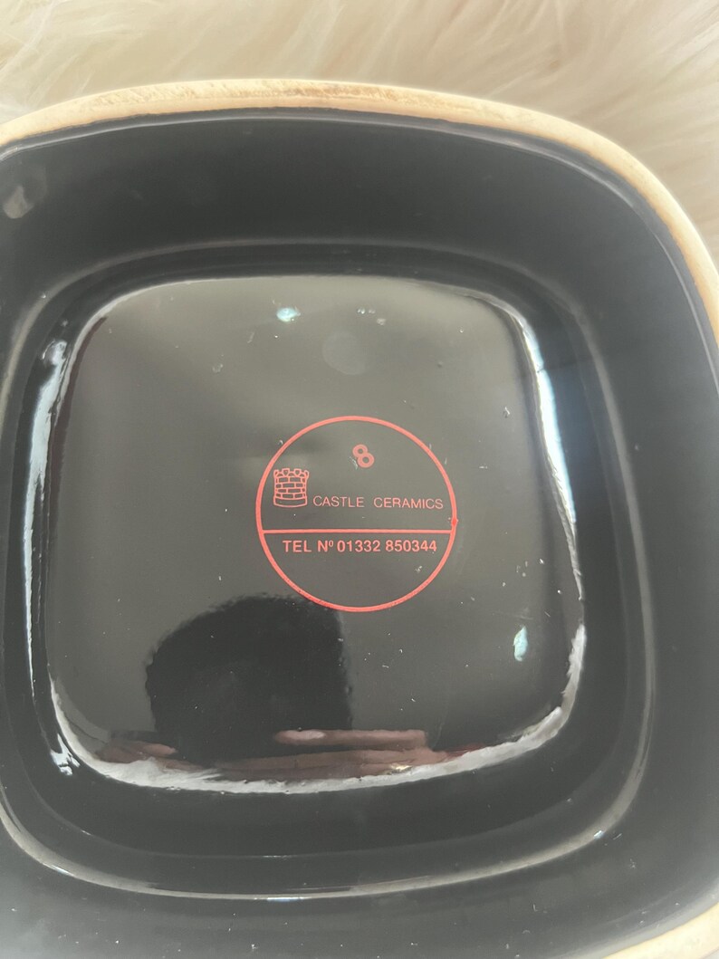 Puede incluir: Un cuenco cuadrado de cer&aacute;mica con interior negro brillante y borde exterior beige claro. Una pegatina roja circular est&aacute; centrada en la base, con un icono de castillo, el texto CASTLE CERAMICS, el n&uacute;mero 8 y el tel&eacute;fono TEL N&deg; 01332 850344.