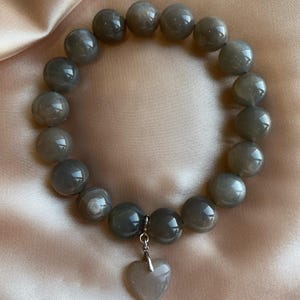 Könnte beinhalten: Ein Armband aus grauen Edelsteinen mit einem herzförmigen Anhänger. Das Armband besteht aus runden Perlen und hat einen silbernen Anhänger.