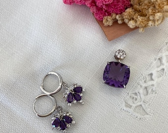 May include: A pair of silver hoop earrings with dangling purple gemstone accents and a single pendant featuring a large, square-shaped purple gemstone with silver accents.