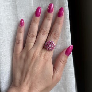 Op de afbeelding: Een hand met een grote, ronde ring met een cluster van roze edelstenen en kleine, heldere stenen. De ring is het middelpunt, tegen een achtergrond van witte stof. De nagels zijn felroze geverfd met witte glitteraccenten.