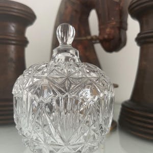 Op de afbeelding: Een glazen snoepbakje met deksel voorzien van een gefacetteerd ster- en ruitpatroon. De houder heeft een ronde vorm en een kleine, gladde knop als handvat aan de bovenzijde, met ingewikkelde geometrische slijpingen die het licht reflecteren.