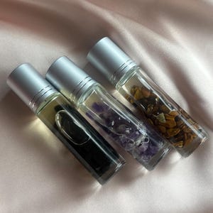 Könnte beinhalten: Drei Glas-Rollerflaschen für ätherische Öle mit silbernen Kappen. Die Flaschen enthalten unterschiedlich farbige Inhalte: schwarze Flüssigkeit, violette Steine und braune Steine. Die Flaschen sind auf einer rosa Satin-Oberfläche angeordnet.