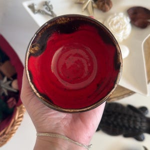 Könnte beinhalten: Eine handgefertigte Keramikschale mit leuchtend rotem Innenraum und dunkelbraunem Rand. Die Außenseite der Schale hat einen natürlichen, erdigen Farbton. Die Schale wird in einer Hand gehalten, wodurch ihre Form hervorgehoben wird. Ideal zum Servieren oder Dekorieren.