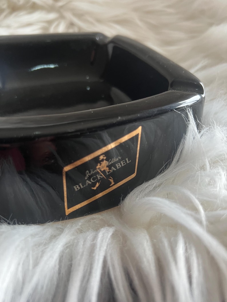 Puede incluir: Un cenicero cuadrado de color negro brillante con un emblema trapezoidal dorado en la parte frontal. El emblema muestra el texto Johnnie Walker BLACK LABEL en una fuente con serifa, acompa&ntilde;ado de un peque&ntilde;o gr&aacute;fico dorado de una figura.