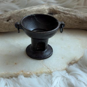 Puede incluir: Un pequeño recipiente de hierro fundido oscuro con una parte superior ancha en forma de cuenco, un cuello cilíndrico estrecho y una base circular. El recipiente cuenta con dos pequeñas asas de anillo integradas en los lados opuestos del borde.