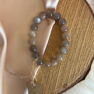 Könnte beinhalten: Ein Armband aus hellbraunen und grauen Sunstone-Perlen. Das Armband liegt auf einer Holzoberfläche mit einem Stück braunem Stoff.