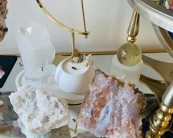 May include: A collection of crystals and geodes on a gold stand, including a white geode, a clear quartz crystal, a pink and white agate, and a white candle with a floral accent.