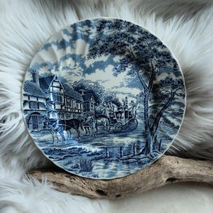 Può includere: Un piatto rotondo in ceramica con un motivo decorativo blu e bianco. Il design raffigura una scena storica con una carrozza trainata da cavalli, edifici a graticcio, un grande albero e uno specchio d'acqua, con un bordo smerlato.
