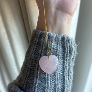 Könnte beinhalten: Eine goldene Halskette mit einem herzförmigen, hellrosa Anhänger. Der Anhänger besteht aus poliertem Stein. Die Halskette wird vor einem grauen Strickpullover gezeigt.