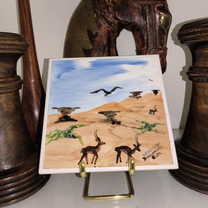 May include: A square ceramic tile featuring a hand-painted desert landscape with tan sand dunes, a blue sky, and several gazelles. The artwork includes green desert vegetation, a bird in flight, and the text "GEARTH/90 DIOLORIPPS RITZ" in the corner.
