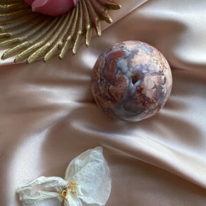 Op de afbeelding: Een roze roosvormige kaars en een decoratief goudkleurig metalen stuk zijn gepositioneerd in de buurt van een gemarmerde bol met een gat. Er is ook een gedroogde witte bloem aanwezig. De items rusten op een satijnen stof.