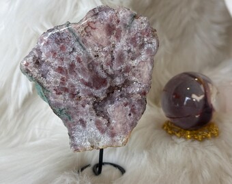May include: A large, pink and green amethyst geode on a black metal stand. The geode has a rough, natural texture and is filled with crystals.