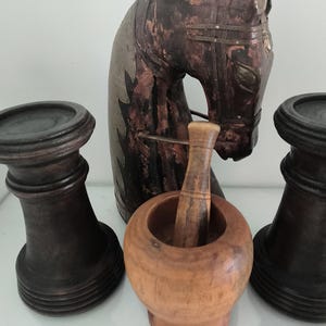 Könnte beinhalten: Eine dunkelbraune hölzerne Pferdekopfskulptur hinter einem hellbraunen hölzernen Mörser mit Stößel, flankiert von zwei dunkelbraunen hölzernen Säulenkerzenhaltern mit kreisförmigen Sockeln und abgestuften Oberteilen, auf einer weißen Fläche.
