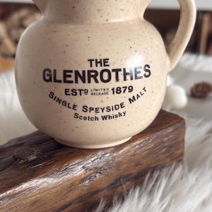 May include: A speckled beige ceramic pitcher with a handle, bearing the text "THE GLENROTHES" and "SINGLE SPEYSIDE MALT SCOTCH WHISKY". The pitcher rests on a wooden block, set against a fluffy white backdrop.