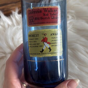 May include: A blue glass with Johnnie Walker Red Label Old Scotch Whisky labels. The labels list awards from Sydney, Paris, Adelaide, and Melbourne. The glass is held by a person.