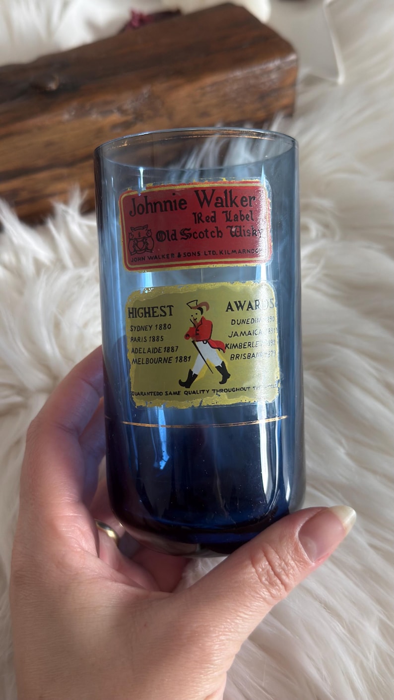 May include: A blue glass with Johnnie Walker Red Label Old Scotch Whisky labels. The labels list awards from Sydney, Paris, Adelaide, and Melbourne. The glass is held by a person.