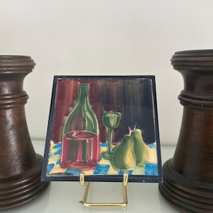May include: A square ceramic tile featuring a painted still life of a green bottle, a red glass, a green stemmed glass, and two green pears on a blue and white checkered tablecloth, displayed on a gold metal stand between two dark wood pillars.