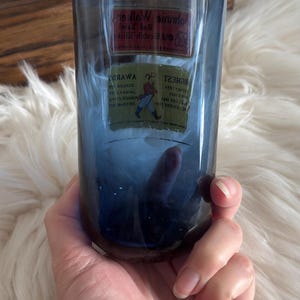 May include: A blue-tinted glass tumbler with a rectangular label featuring text and a logo. The glass is held in a hand against a soft, white background. The tumbler appears to be a vintage item.