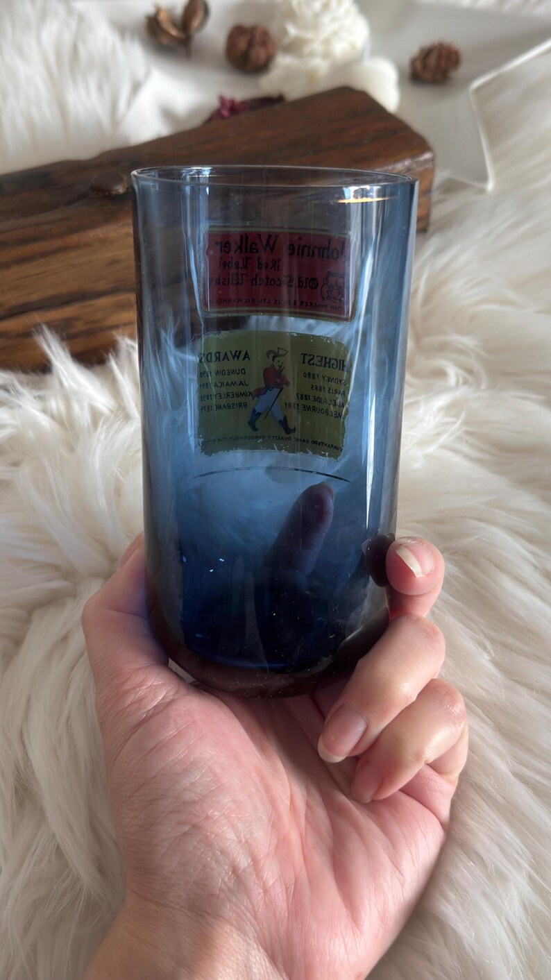 May include: A blue-tinted glass tumbler with a rectangular label featuring text and a logo. The glass is held in a hand against a soft, white background. The tumbler appears to be a vintage item.
