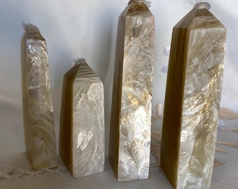 May include: Four iridescent white and gold crystal points, also known as selenite, arranged in a row on a white surface.