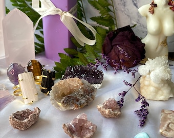 May include: A collection of amethyst, rose quartz, and other crystals, including a purple candle, a white candle in the shape of a torso, and a few rings. The crystals are arranged on a white surface with a green fern in the background.