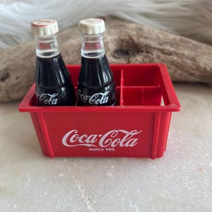 以下が含まれることがあります： コカ・コーラのロゴとMARCA REGの文字が記された赤いプラスチック製のミニチュアケース。中には、暗い液体が入った白いキャップ付きのガラスのような小さなボトルが2本収められています。ボトルにもコカ・コーラのロゴが印字されています。