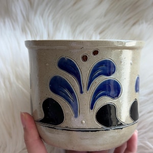 May include: A cylindrical ceramic crock with a light beige glazed finish. The exterior features a hand-painted pattern of cobalt blue stylized leaf motifs and black curved accents, complemented by two small brown dots near the top rim.