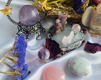 May include: An assortment of crystals and decorative items. A large amethyst crystal ball sits in a silver stand with a gold star and crescent moon. Other items include a pink heart-shaped stone, a small crystal ball, and a white stone with dark spots.