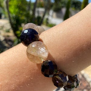 Könnte beinhalten: Ein Armband mit facettierten, runden Perlen in Schwarz, Braun und Klar mit goldenen Einschlüssen. Das Armband wird am Handgelenk getragen, mit einem verschwommenen Hintergrund aus grünem Laub und blauem Himmel.