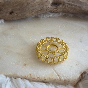 May include: A circular gold-toned metal finding featuring a delicate filigree pattern with heart-shaped cutouts and a central circular setting. The component has a textured, beaded edge and a raised rim, designed for jewellery making and crafting projects.