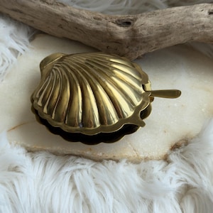 May include: A polished gold-toned brass shell-shaped container with a hinged lid and a small matching spoon extending from the side. The metal surface features prominent vertical ridges and a smooth, reflective finish, capturing light across its curved form.